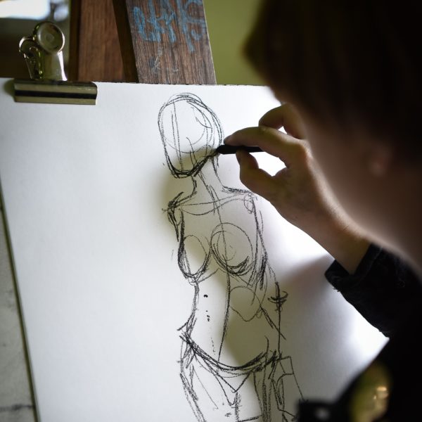 A hand holding charcoal, drawing a figure of a nude woman